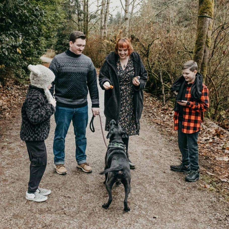 A man, woman and two pre-teen children stand on a woodland path facing their dog with treats in their hands. The dog is an adult black lab.