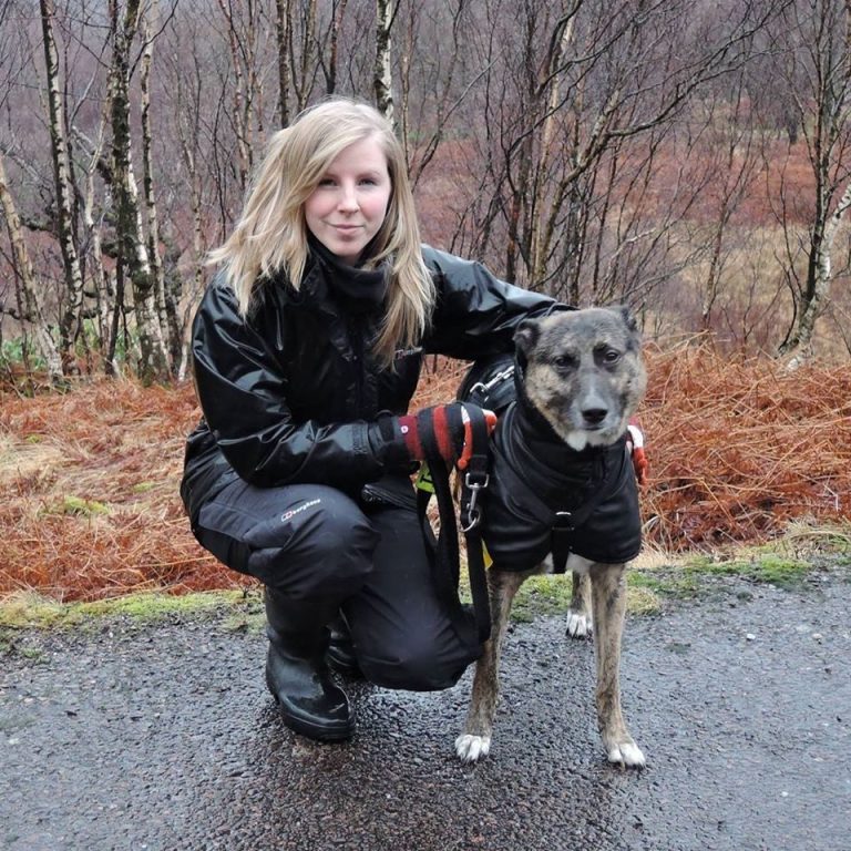 A white woman in her late 20s, long blonde hair, wearing black waterproofs crouched by a brindle Lurcher type wearing a black raincoat.